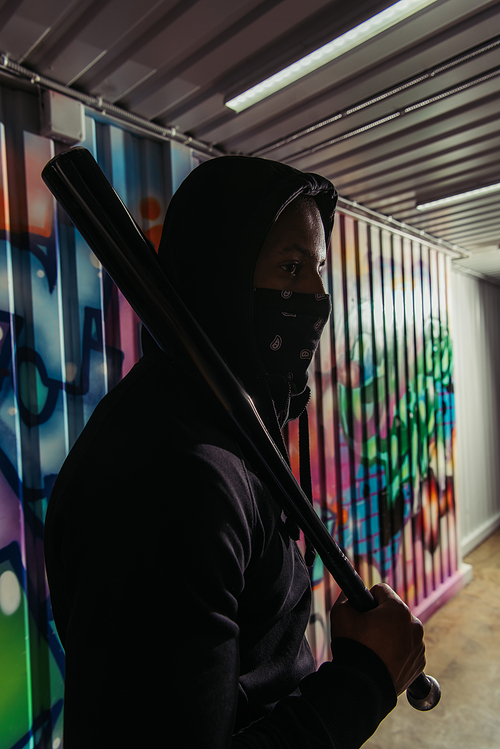 African american hooligan in mask holding baseball bat near graffiti