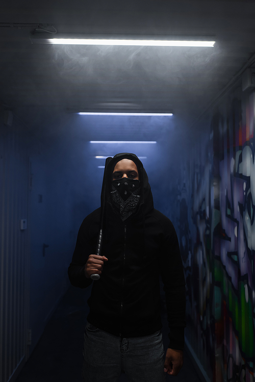 African american hooligan in mask holding baseball bat and looking at camera near lighting and graffiti