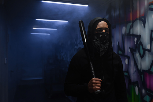 African american hooligan holding baseball bat near graffiti and smoke in garage