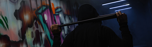 Back view of african american bandit in hoodie holding baseball bat in garage with graffiti, banner