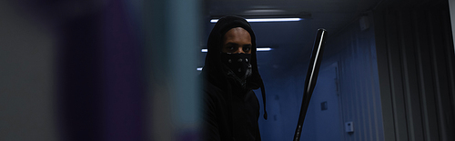 African american hooligan with mask on face holding baseball bat in garage, banner