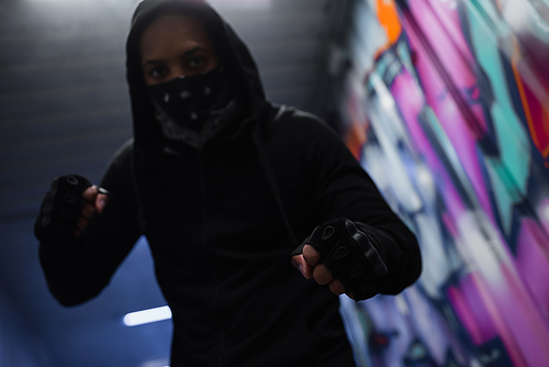 Low angle view of blurred african american hooligan in gloves and mask standing in fighting pose in garage