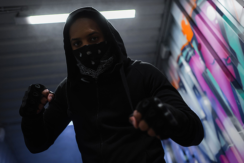 Low angle view of african american bandit in mask standing in fighting pose near graffiti in garage