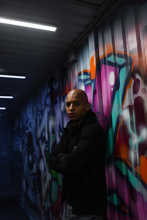 African american hooligan crossing arms near graffiti on wall in garage