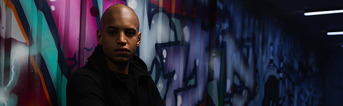 African american hooligan looking at camera near graffiti on wall in garage, banner