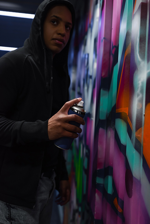 Blurred african american vandal in hood painting graffiti and looking away in garage