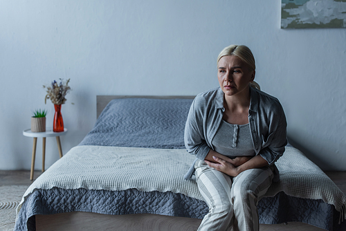 upset blonde woman with menopause suffering from stomach ache in bedroom