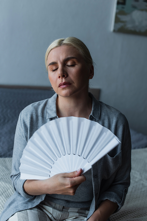 displeased blonde woman with menopause suffering from heat and cooling with fan in bedroom