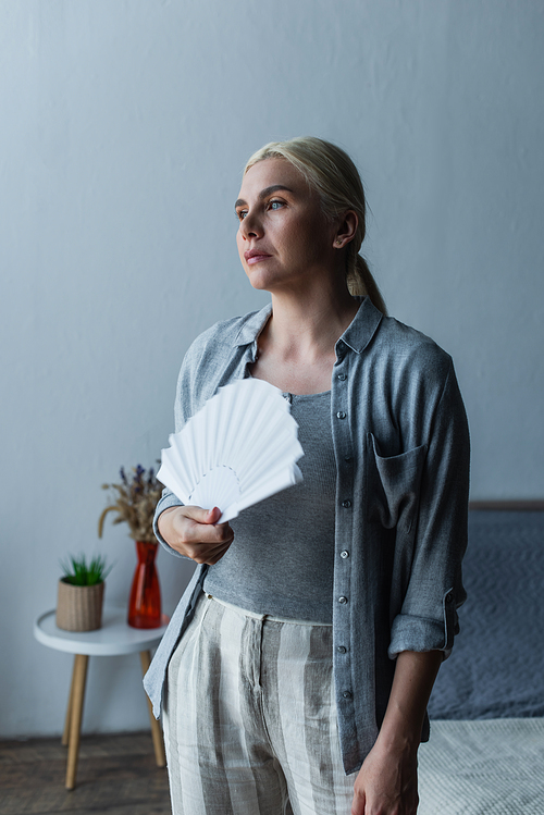 blonde woman with menopause suffering from heat and holding fan in bedroom
