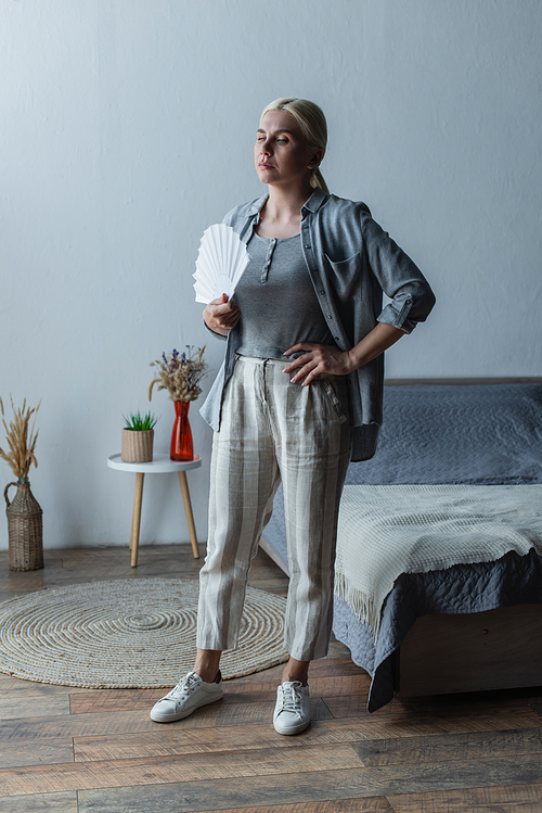 full length of blonde woman with menopause suffering from heat and holding fan in bedroom