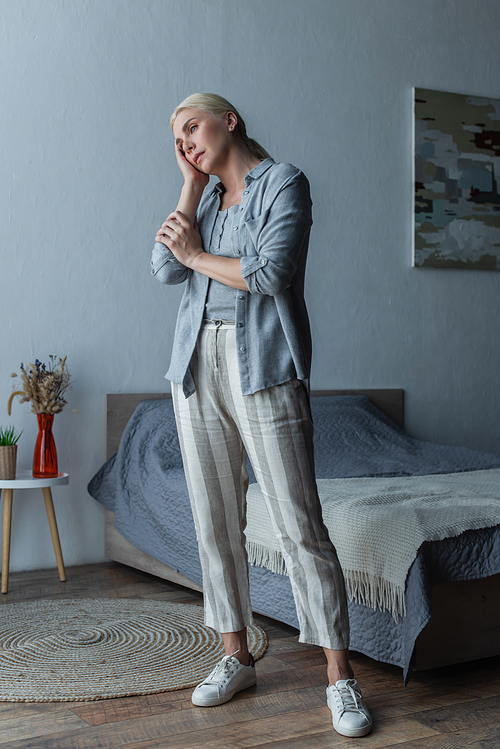 full length of tired woman with menopause standing and touching forehead in bedroom