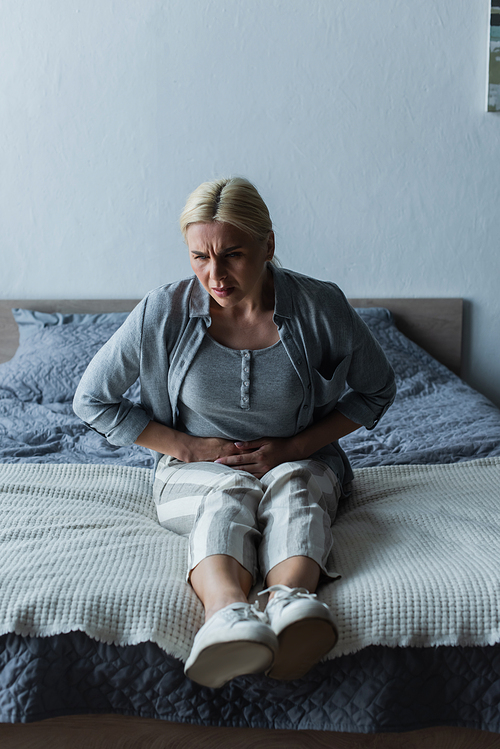 upset blonde woman with menopause suffering from abdominal pain in bedroom
