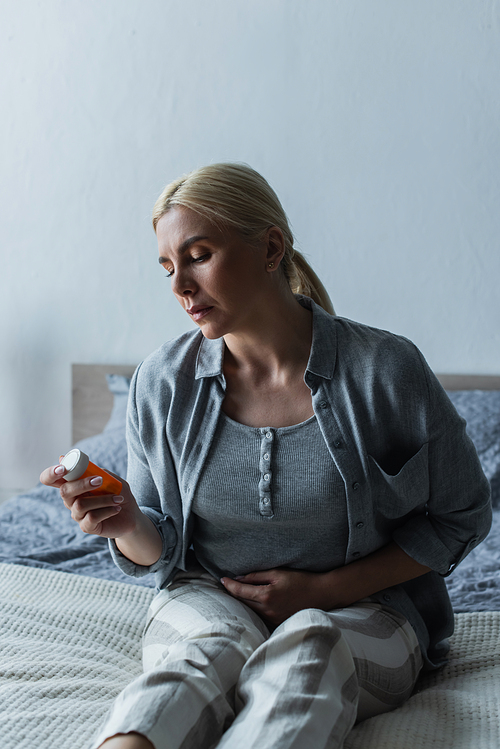 depressed blonde woman with menopause holding bottle with pills and sitting on bed
