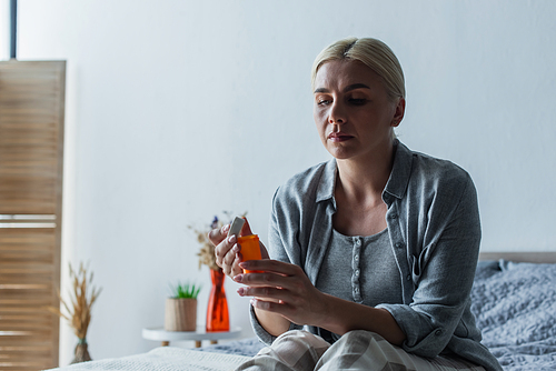 tired blonde woman with menopause holding bottle with pills and sitting on bed