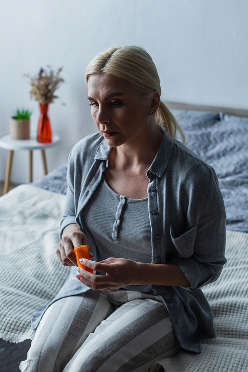 upset woman with menopause holding bottle with medication and sitting on bed