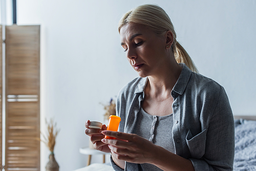 frustrated blonde woman with menopause holding bottle with pills and sitting in bedroom
