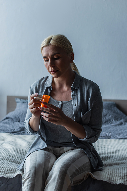 sad blonde woman with menopause holding bottle with pills and sitting on bed