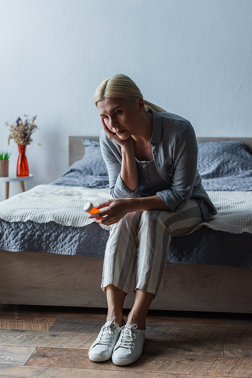 upset blonde woman with menopause holding bottle with pills and sitting on bed