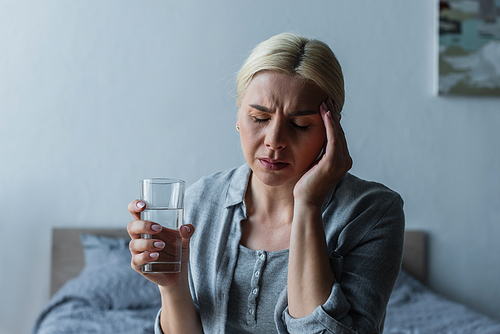 blonde woman with menopause suffering from headache and holding glass of water