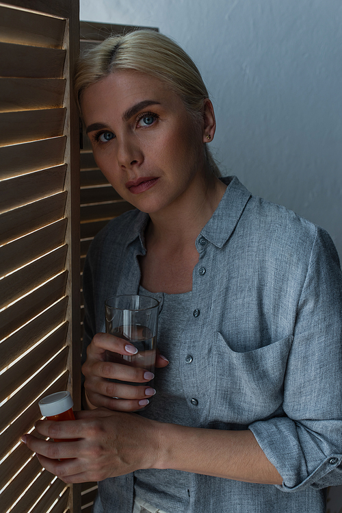 blonde woman with menopause holding glass of water near folding screen