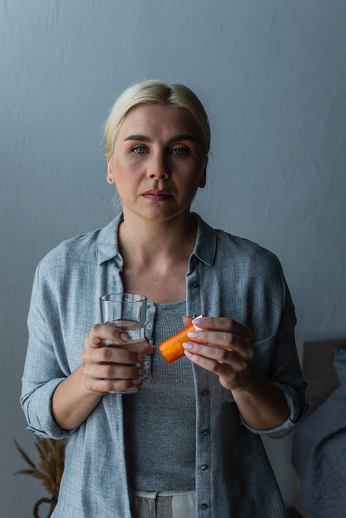 blonde woman with menopause holding glass of water and bottle with medication