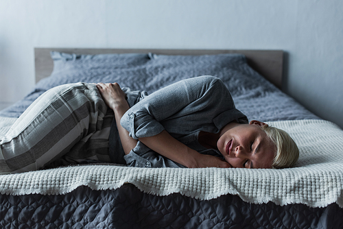 blonde woman lying on bed and suffering from stomach ache during menopause