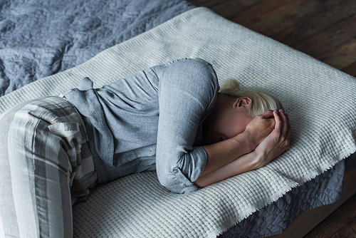 blonde woman lying on bed and covering face while suffering from stomach ache during menopause