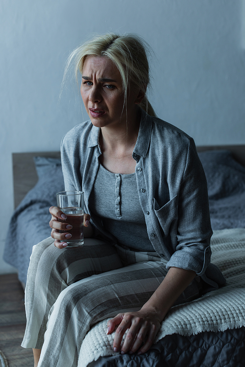 exhausted woman holding glass of water while sitting on bed during menopause