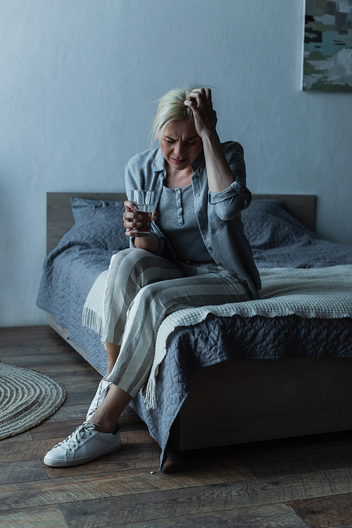 full length of exhausted woman holding glass of water while having migraine during menopause