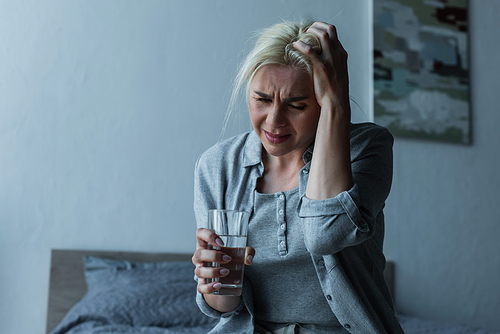 exhausted blonde woman holding glass of water while having migraine during menopause