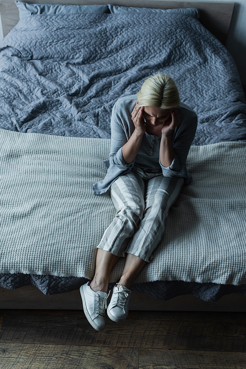 high angle view of blonde woman sitting on bed while having headache during menopause