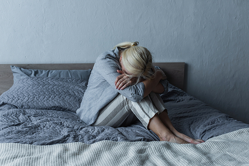 blonde woman covering face and hugging knees on bed while feeling pain in stomach during menopause