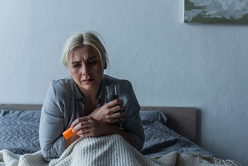 exhausted woman with climax sitting in bed while holding glass of water and painkillers