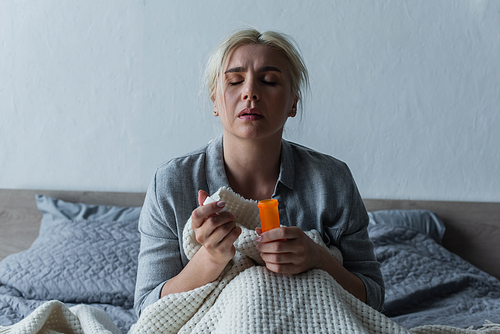 sad woman with climax sitting in bed and holding bottle with painkillers