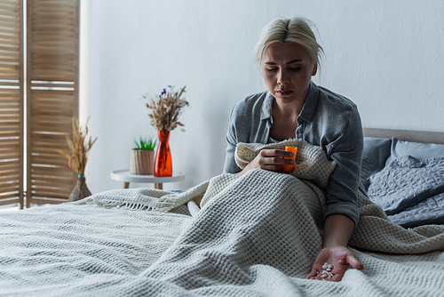 sad woman with climax looking at bottle with painkillers while sitting in bed under blanket