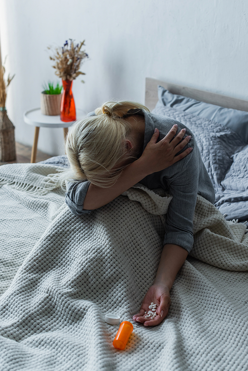 blonde woman with climax covering face while sitting in bed with painkillers in hand