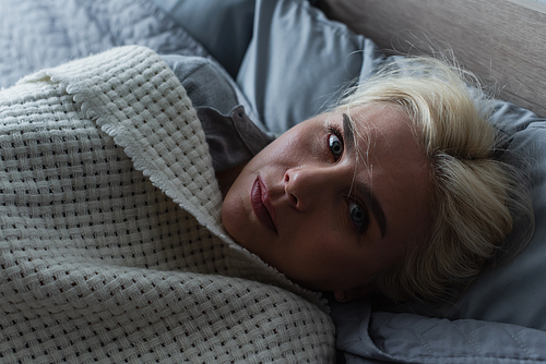 top view of depressed blonde woman with menopause lying under blanket in bed