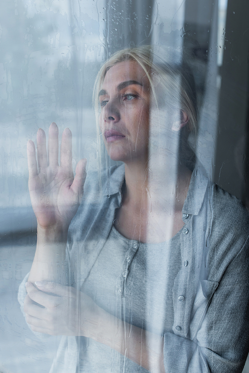 depressed blonde woman looking through wet window with rain drops