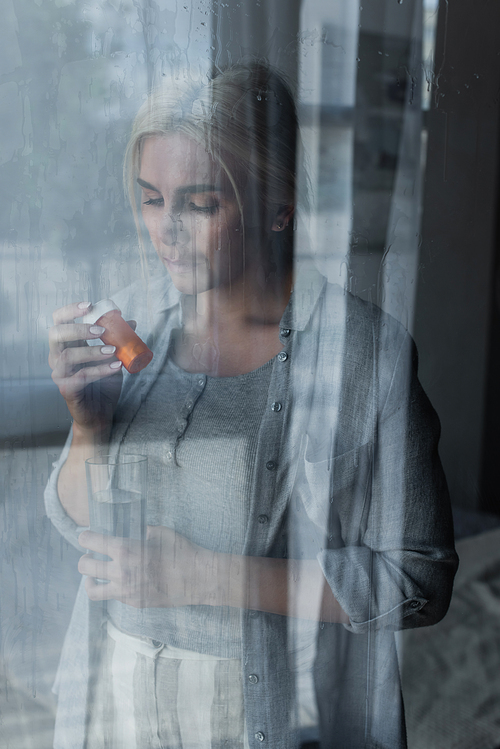 depressed blonde woman holding bottle with pills and glass of water near window with rain drops