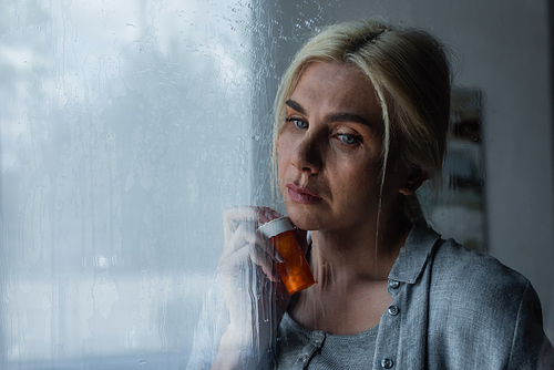 depressed blonde woman holding bottle with pills near window with rain drops