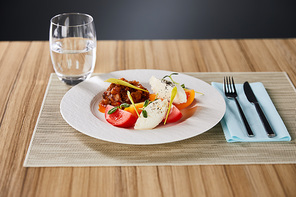selective focus of delicious restaurant dish with eggplant caviar and tomatoes served on wooden table with water and cutlery isolated on grey