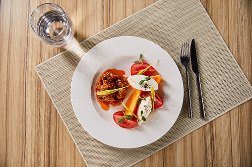 top view of delicious restaurant dish with eggplant caviar and tomatoes served on wooden table with water and cutlery