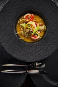 top view of delicious restaurant soup with shrimps in black plate with cutlery