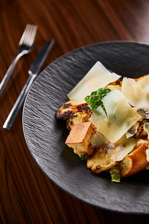 selective focus of tasty Caesar salad with Parmesan slices served on plate with cutlery on wooden table