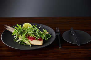 tasty restaurant fish steak with lime and arugula on wooden table near cutlery on black background