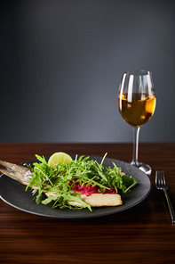 tasty restaurant fish steak with lime and arugula on wooden table near fork and white wine on black background