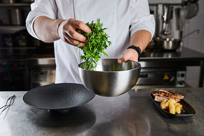 cropped view of chef cooking dish with arugula, meat and potato at kitchen in restaurant