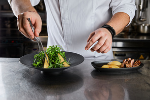 partial view of chef cooking dish with arugula, meat and potato at kitchen in restaurant