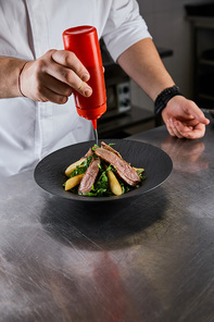 cropped view of chef pouring sauce on dish with arugula, meat and potato at kitchen in restaurant