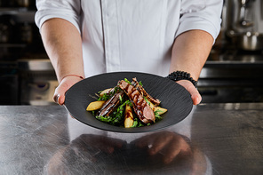 partial view of chef holding dish with arugula, meat and potato at kitchen in restaurant